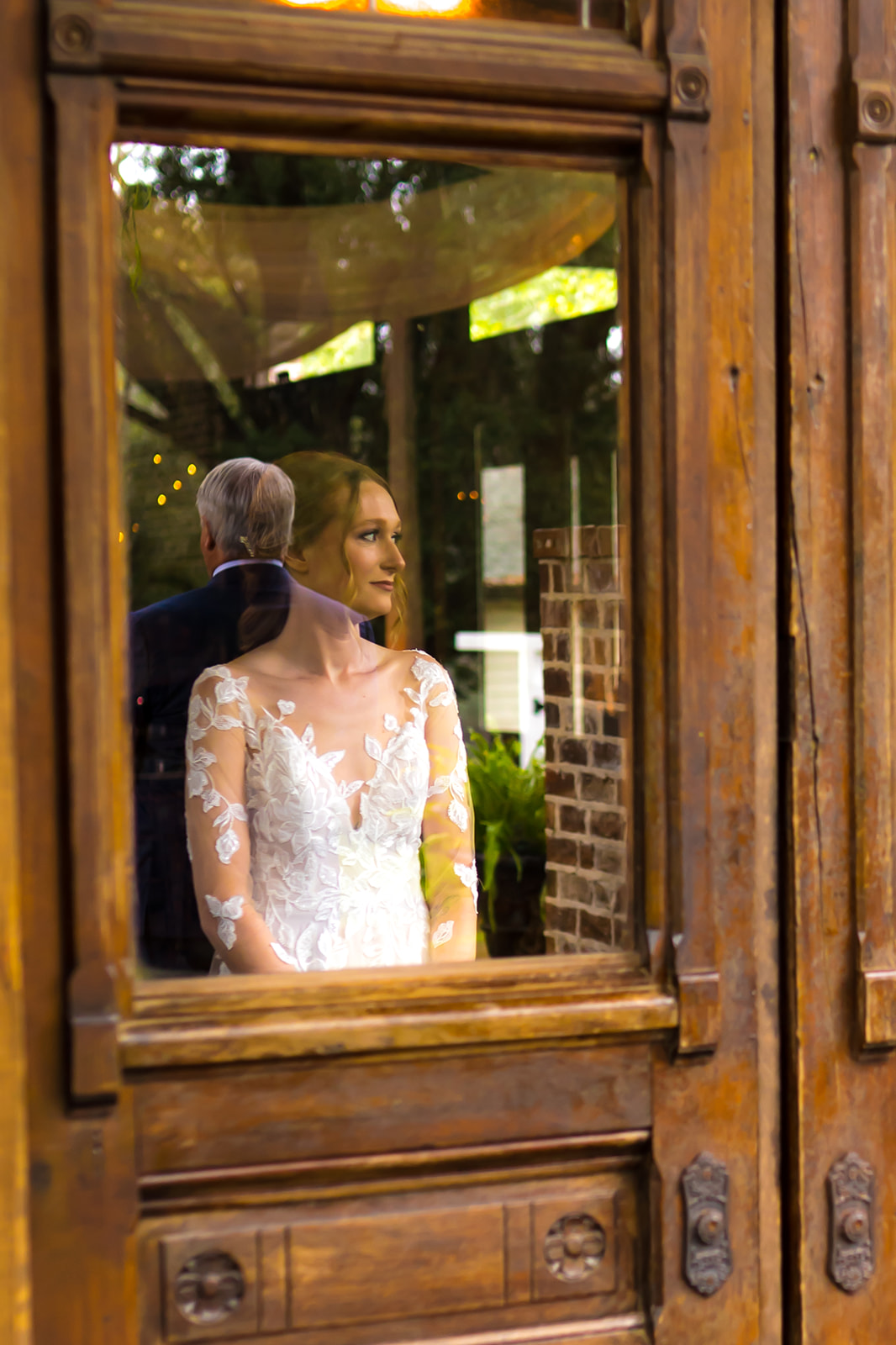 Bride in her white dress looks on with her fathers reflection in a window as she waits for their first look.