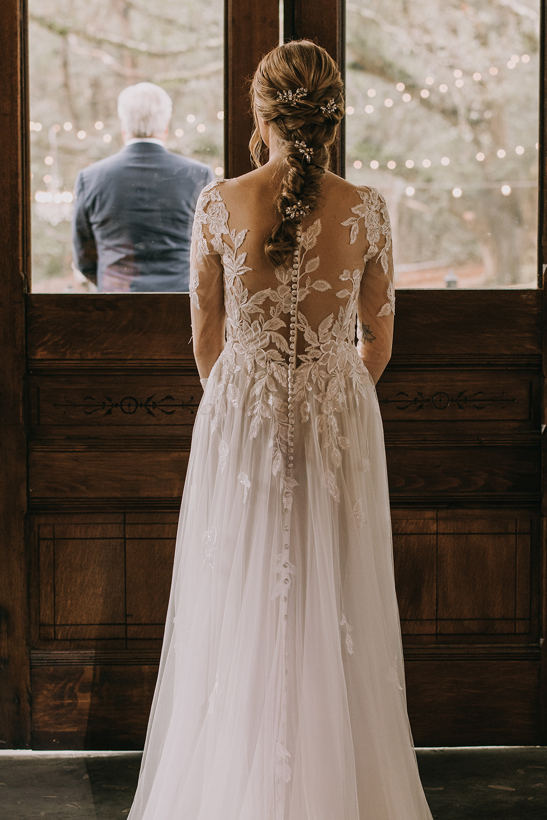 Bride in her white dress looks on through a window before her first look with her father. 