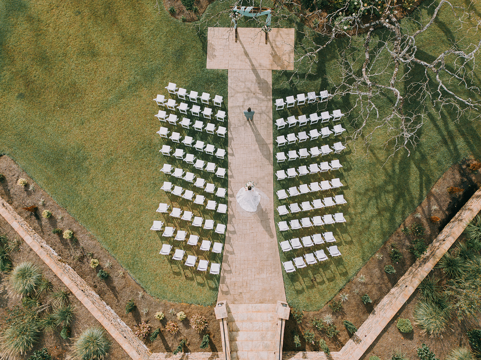 Drone view of a bride and groom during their first look at an outdoor wedding venue in Alabama. The bride, in a flowing white gown, stands on a stone pathway holding her bouquet as she approaches her groom, who is facing away. Rows of white chairs are neatly arranged on either side, leading up to a floral-adorned wedding arch. The setting is surrounded by lush greenery, with tree branches casting dramatic shadows on the grass.